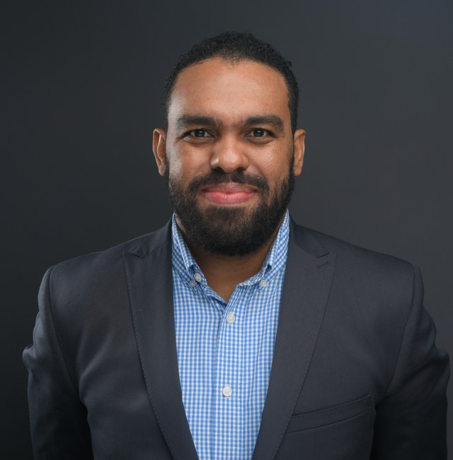 Darwin Santos in a navy blazer and blue checked shirt against a charcoal studio background.